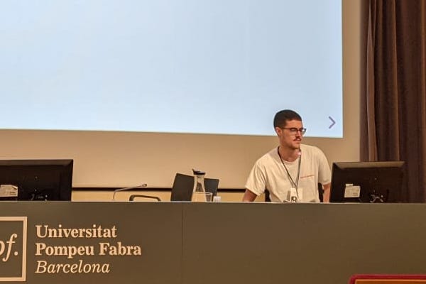A person giving a presentation at an auditorium with a large projection of a title slide in the background